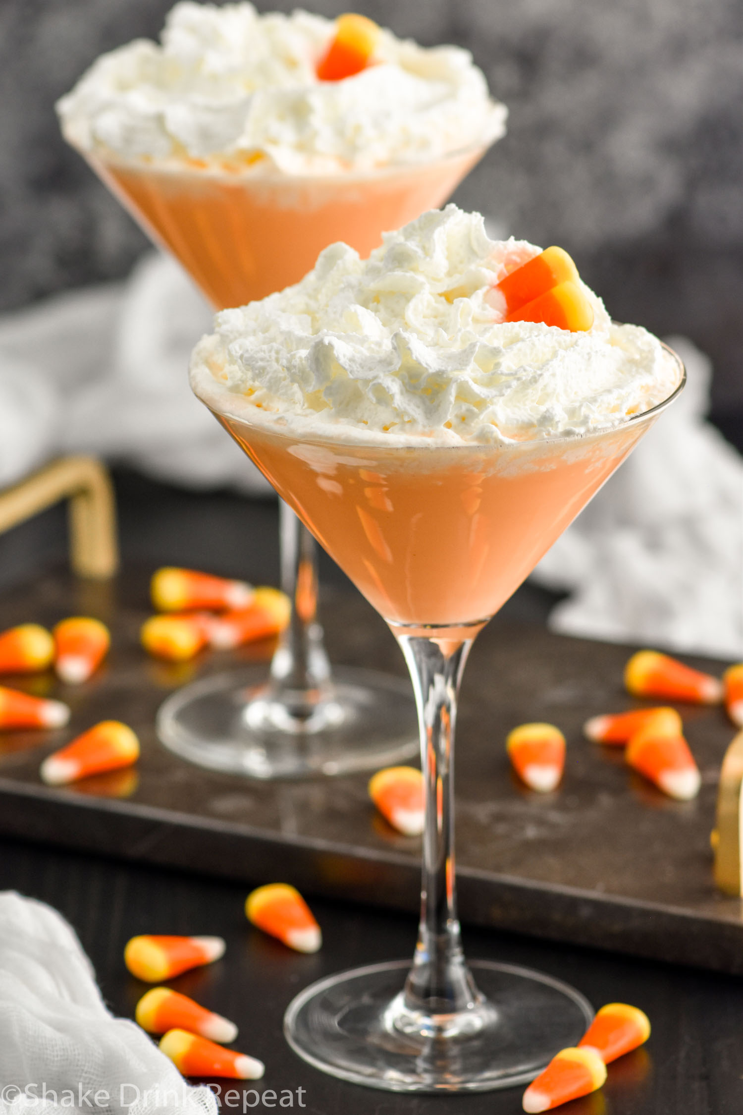 Two martini glasses of candy corn martini topped with whipped cream and garnished with candy corn candy. Candy corn surrounding base of glasses.