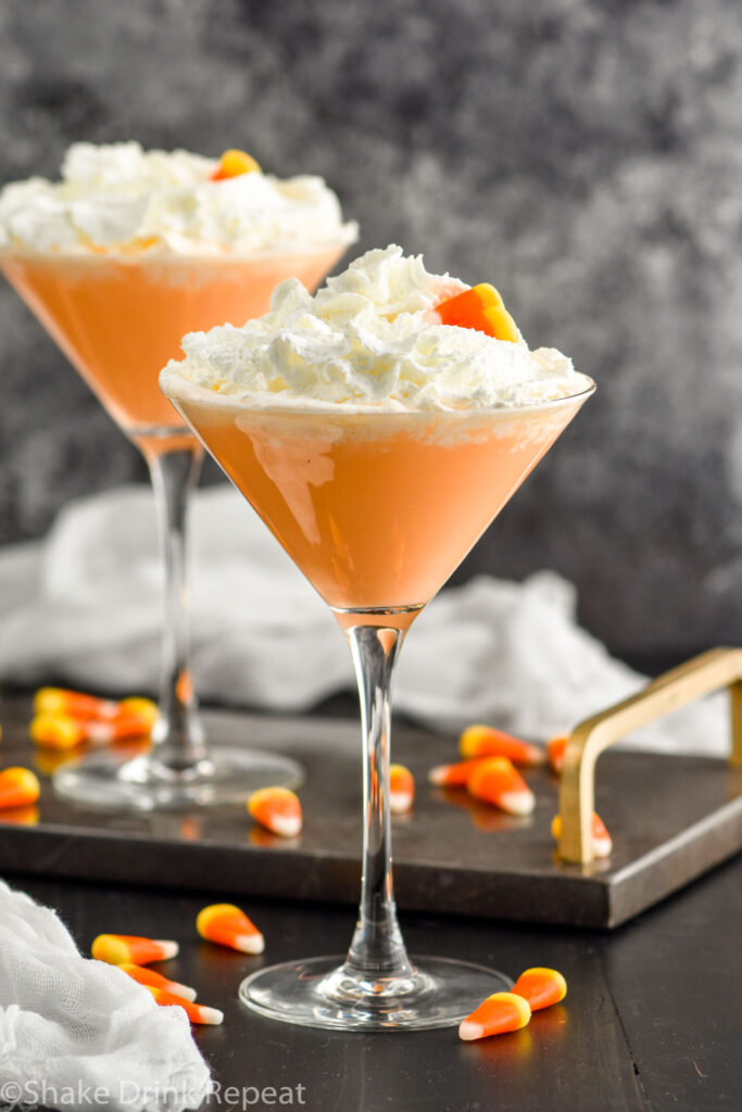Two martini glasses of candy corn martini topped with whipped cream and garnished with candy corn candy. Candy corn surrounding base of glasses.