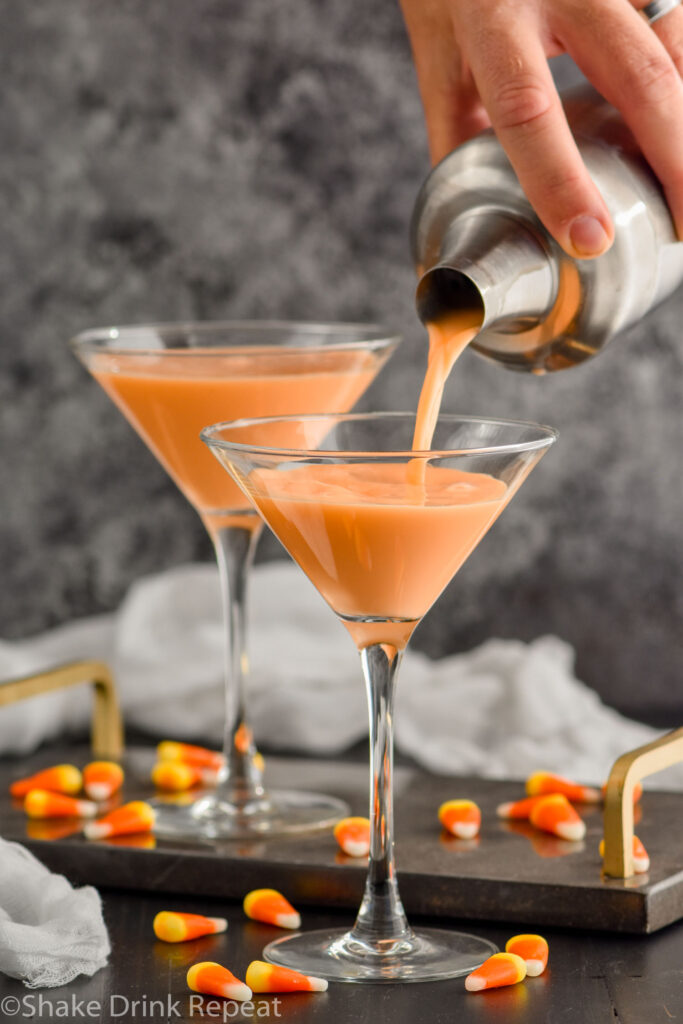 man's hand pouring cocktail shaker of candy corn martini ingredients into a martini glass. Martini glass of candy corn martini recipe sitting in background, candy corn candies surround base of glasses.