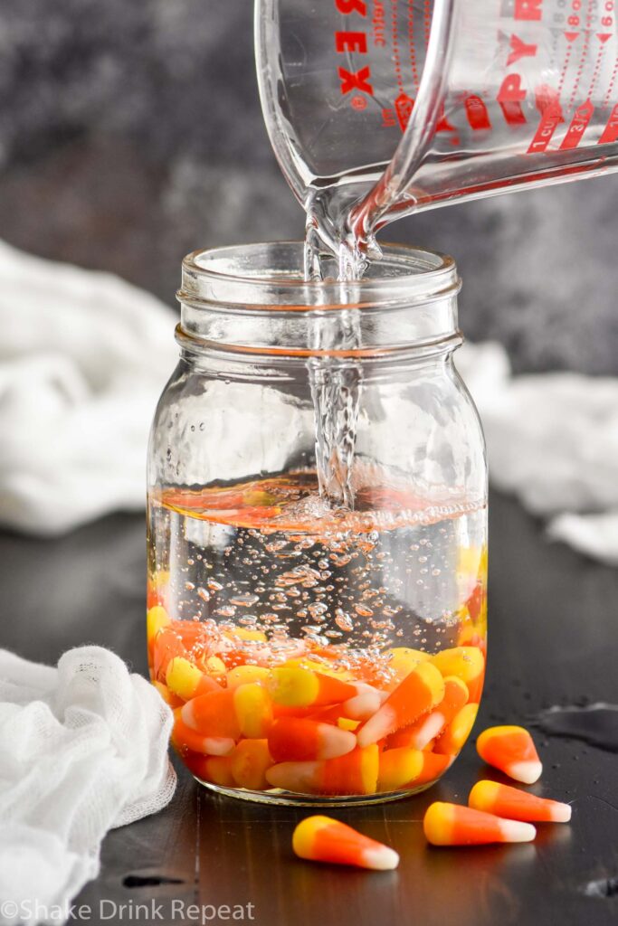 Glass measuring cup pouring vodka into a mason jar of candy corn to make candy corn vodka with candy corns in front of jar.