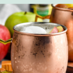 pinterest graphic of caramel apple moscow mules. Text says "caramel apple moscow mules so easy! shakedrinkrepeat.com" Images shows copper mug of caramel apple moscow mule with ice and apple slices surrounded by fresh apples, caramels, and a bottle of ginger beer.