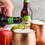 pinterest graphic of caramel apple moscow mules. Text says "the best caramel apple moscow mules only uses 4 simple ingredients! shakedrinkrepeat.com" Image shows man's hand pouring bottle of ginger beer into a copper mug of caramel apple moscow mule ingredients surrounded by caramels, apples, and a bottle of ginger beer.