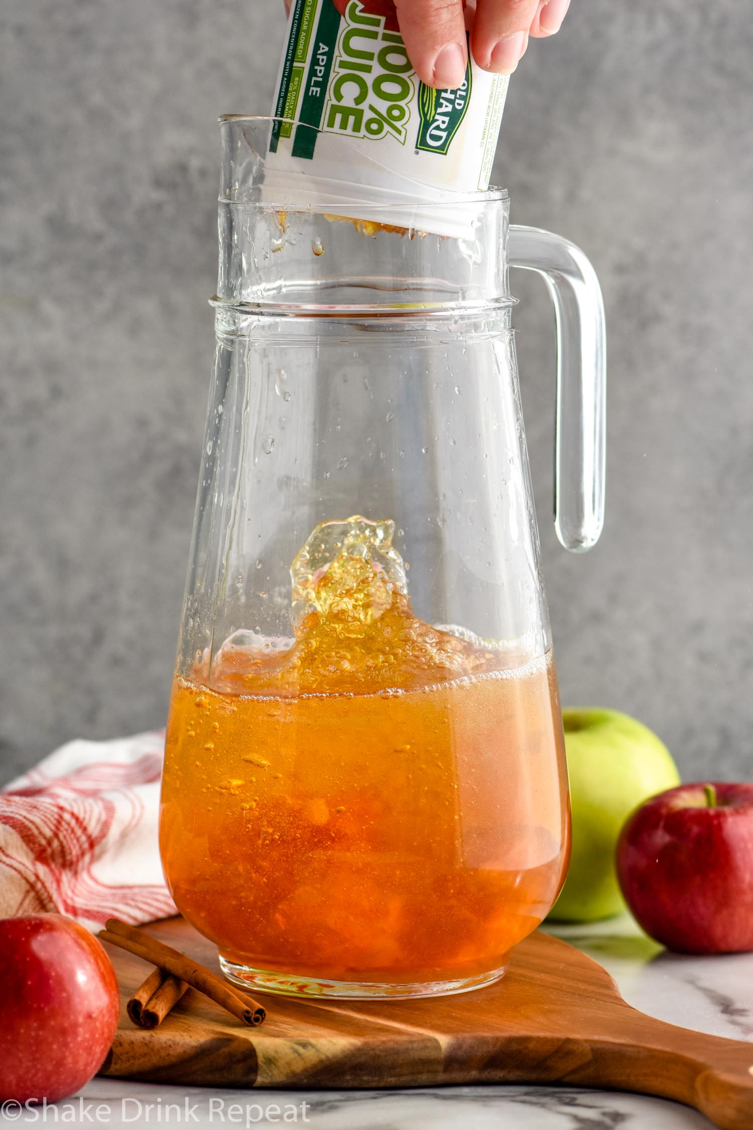 man's hand dumping apple juice concentrate into a pitcher of fireball whiskey punch ingredients surrounded by fresh apples and cinnamon sticks