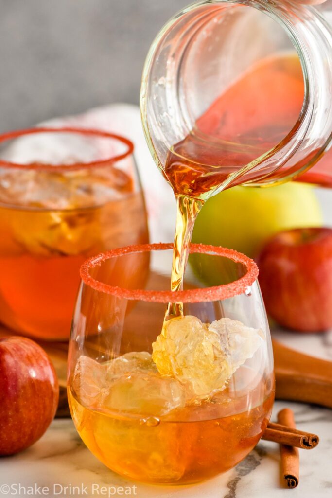 pitcher of fireball whiskey punch pouring into a glass of ice rimmed with red sanding sugar with cinnamon sticks and apples sitting beside