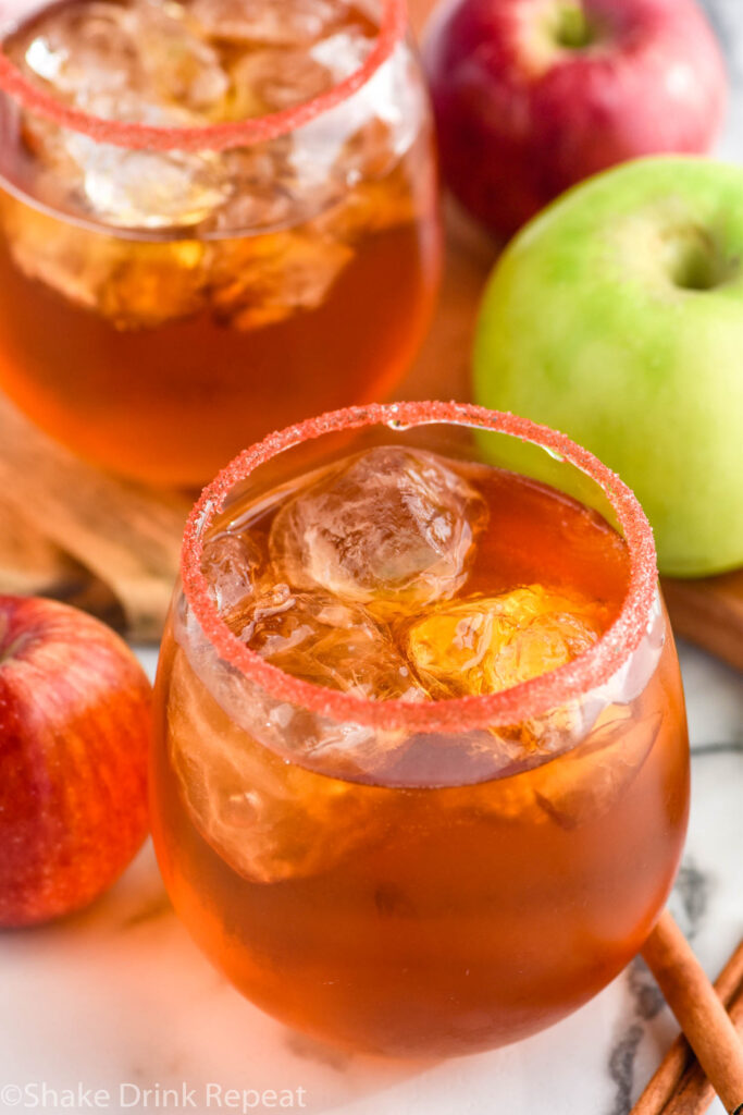 two stemless wine glasses of fireball whiskey punch rimmed with red sugar with ice and fresh apples and cinnamon sticks sitting beside.
