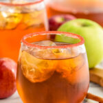 two stemless wine glasses of fireball whiskey punch rimmed with red sugar with ice and fresh apples and cinnamon sticks sitting beside.