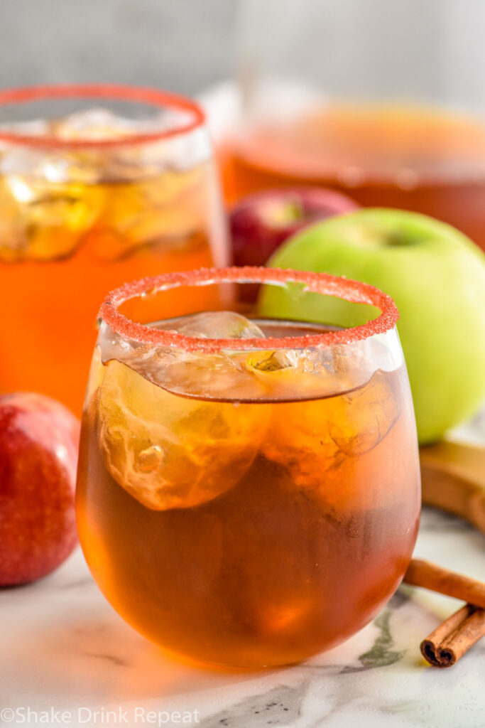 two stemless wine glasses of fireball whiskey punch rimmed with red sugar with ice and fresh apples and cinnamon sticks sitting beside.