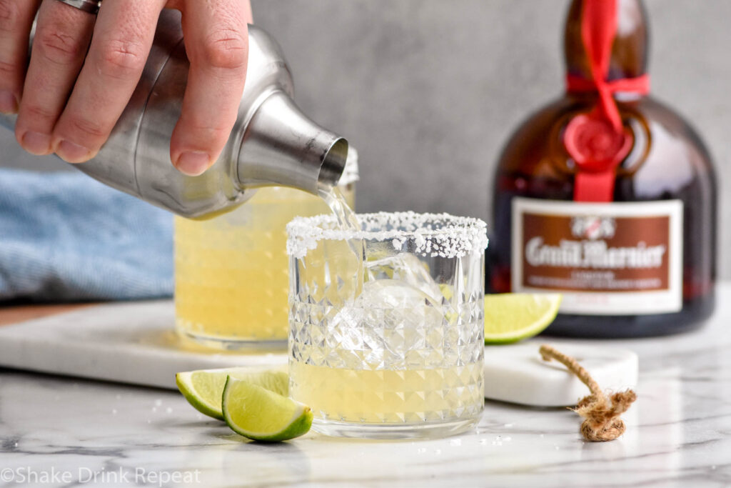 Overhead photo of person's hand pouring a shaker bottle of Grand Marnier Margarita recipe into a glass of ice with a salted rim. Bottle of Grand Marnier liqueur on the counter in the background.