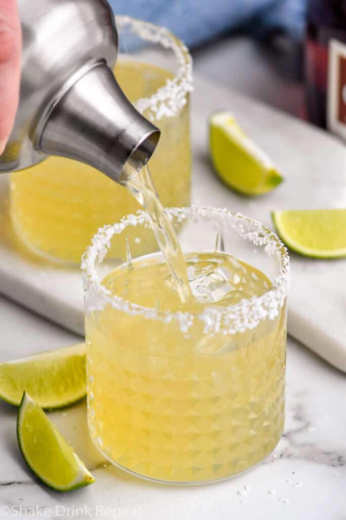 Overhead photo of person's hand pouring a shaker bottle of Grand Marnier Margarita recipe into a glass of ice with a salted rim.