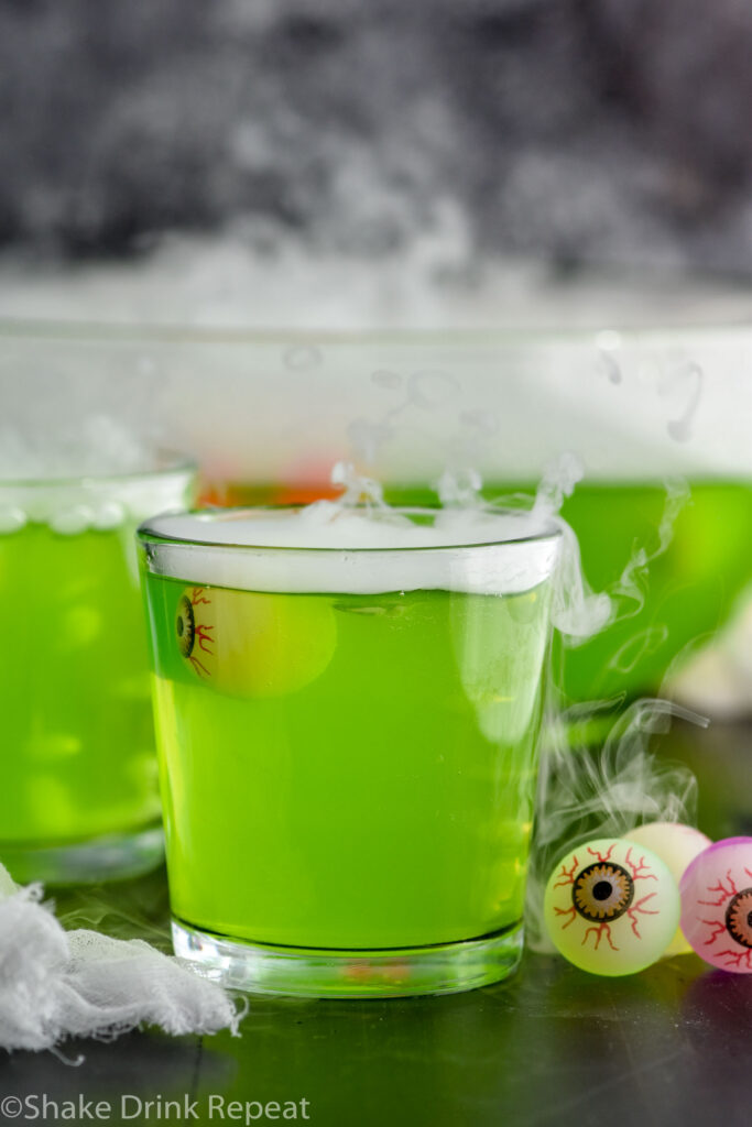 glass of halloween punch topped with dry ice and toy eye balls. Punch bowl of Halloween punch sits in background.