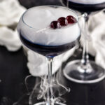 overhead view of two glasses of witches brew cocktails garnished with dark cherries and dry ice