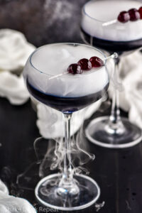 overhead view of two glasses of witches brew cocktails garnished with dark cherries and dry ice