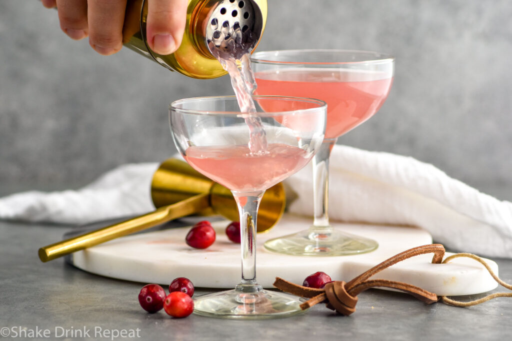 man's hand pouring cocktail shaker of cranberry daiquiri ingredients into a glass with glass of cranberry daiquiri in background and cranberries laying beside glass.