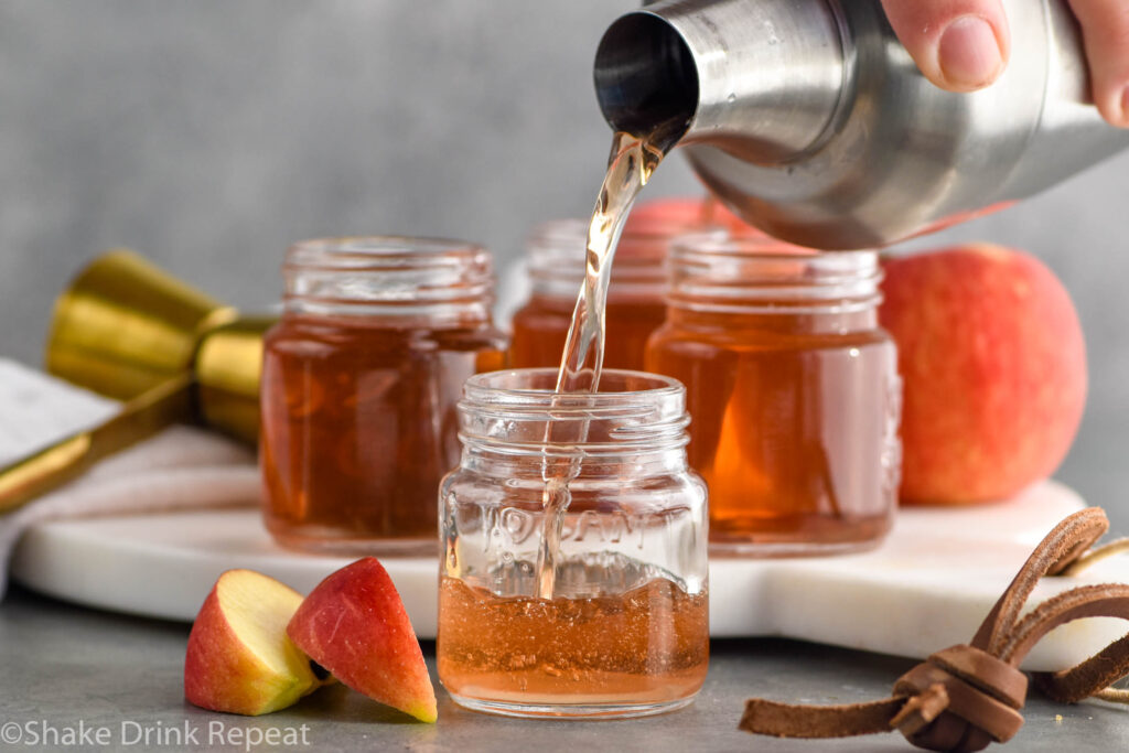 man's hand pouring cocktail shaker of washington apple shot ingredients into a shot glass for serving. Shot glasses of washington apple shots and apple slices sitting in background and beside.