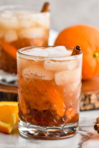 two glasses of fall spiced old fashioned cocktails with ice. Orange slices and cinnamon sticks sitting beside.