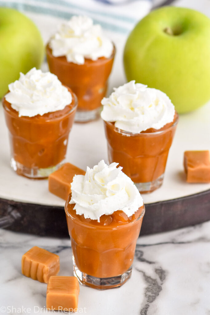 Photo of Caramel Apple Pudding Shots garnished with whipped cream. Apples and caramels sit on counter beside shots.