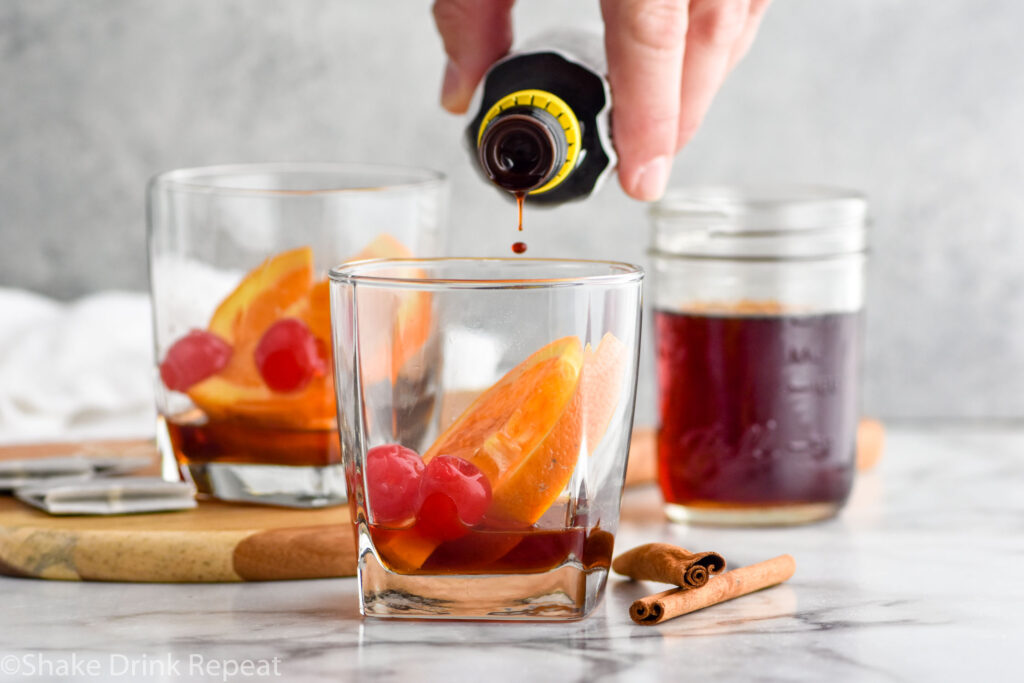 Photo of bitters being added to Chai Old Fashioned recipe. Jar of chai simple syrup on the counter in the background.