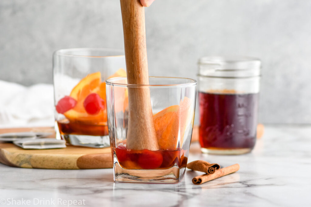 Photo of muddling ingredients for Chai Old Fashioned recipe. Jar of chai simple syrup sits in the background.