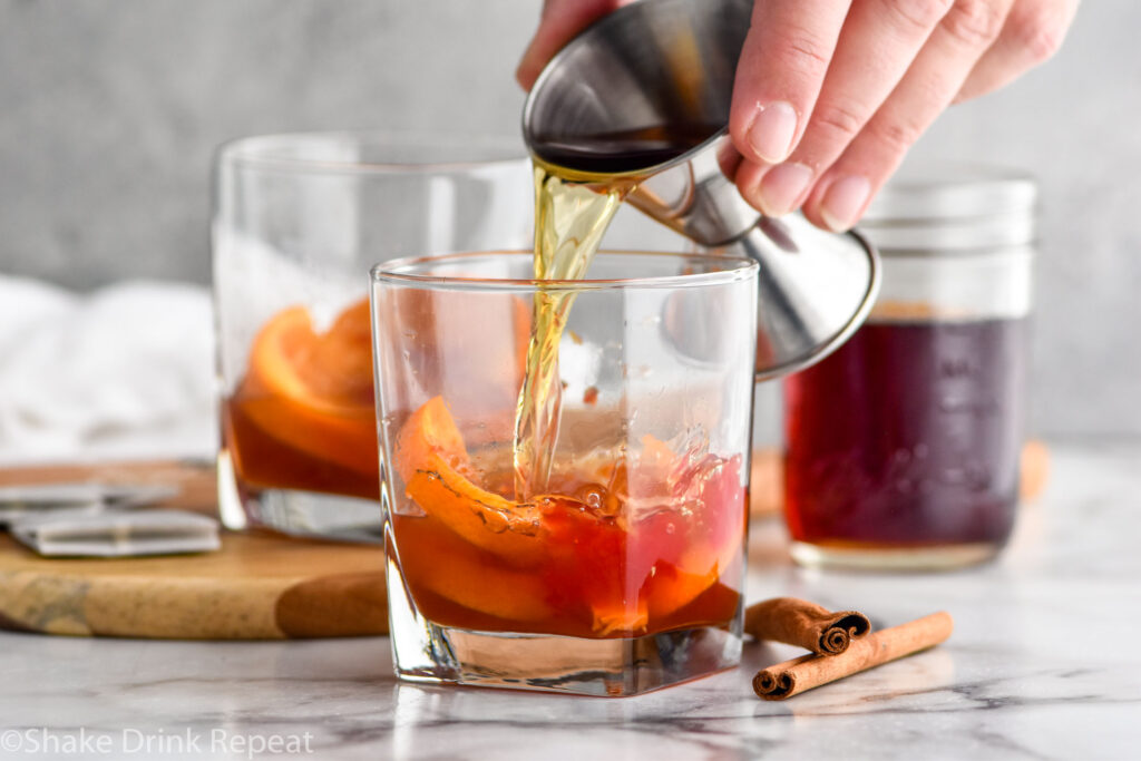 Photo of man's hand pouring bourbon into Chai Old Fashioned recipe. Jar of chai simple syrup is in the background.