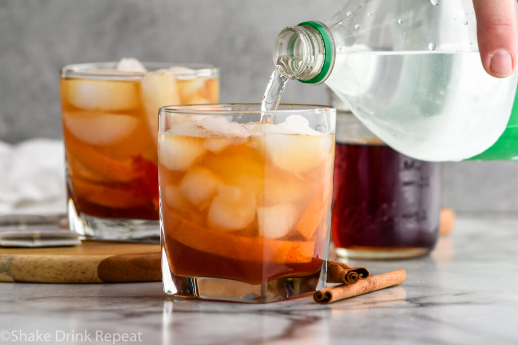 Photo of Chai Old fashioned recipe with man's hand pouring lemon lime soda into cocktail. Jar of chai simple syrup is on the counter.