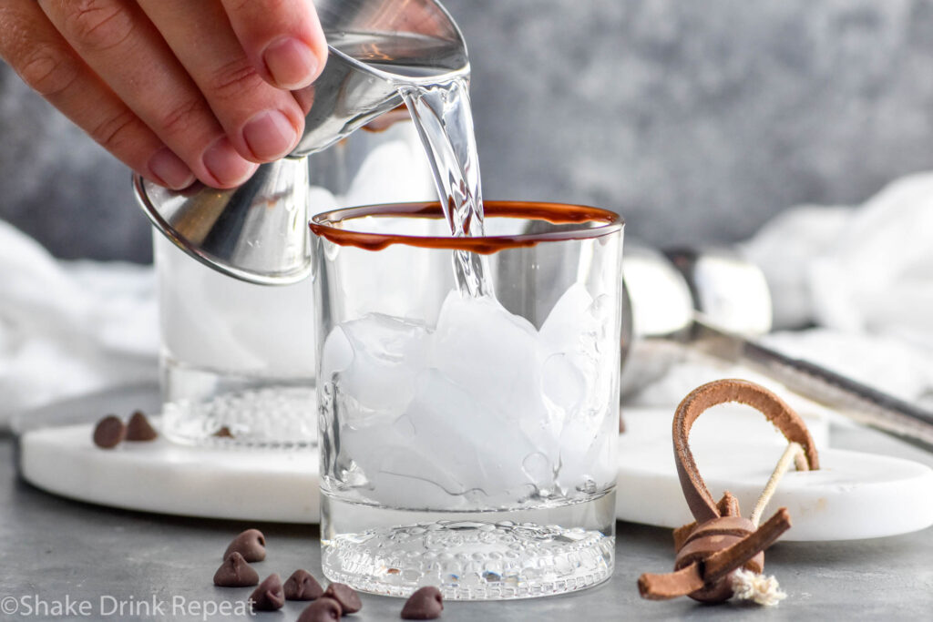 Photo of person's hand pouring vodka into a tumbler of ice with a chocolate rim for chocolate white russian recipe.