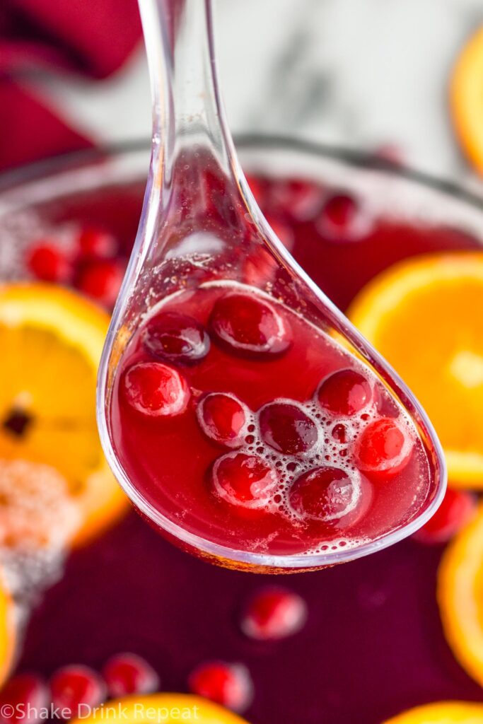 ladle holding Christmas punch with fresh cranberries. Bowl of Christmas punch in background garnished with orange slices and cranberries