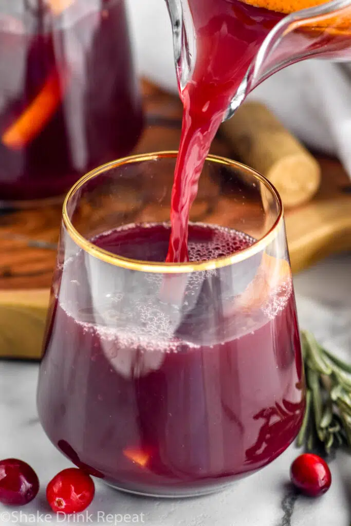 pitcher of cranberry orange sangria pouring into a glass with cranberries and rosemary sprigs sitting beside