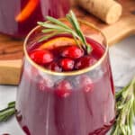 glass of cranberry orange sangria garnished with cranberries and a rosemary sprig with extra cranberries and rosemary sitting beisde
