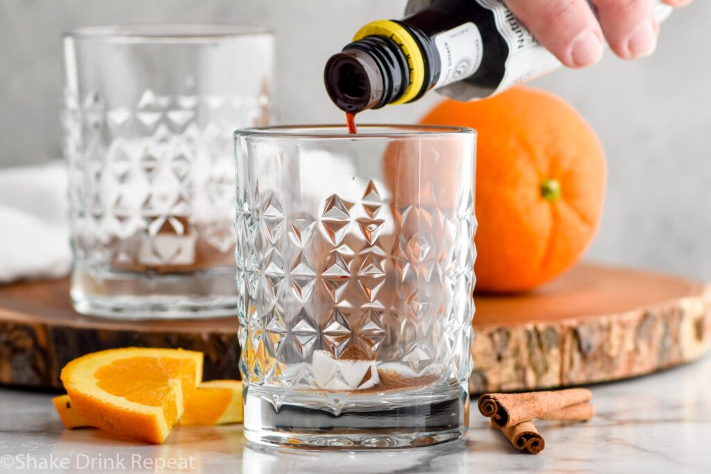 man's hand pouring bitters into a glass with sugar to make a fall spiced old fashioned cocktail with orange slices and cinnamon sticks sitting beside.