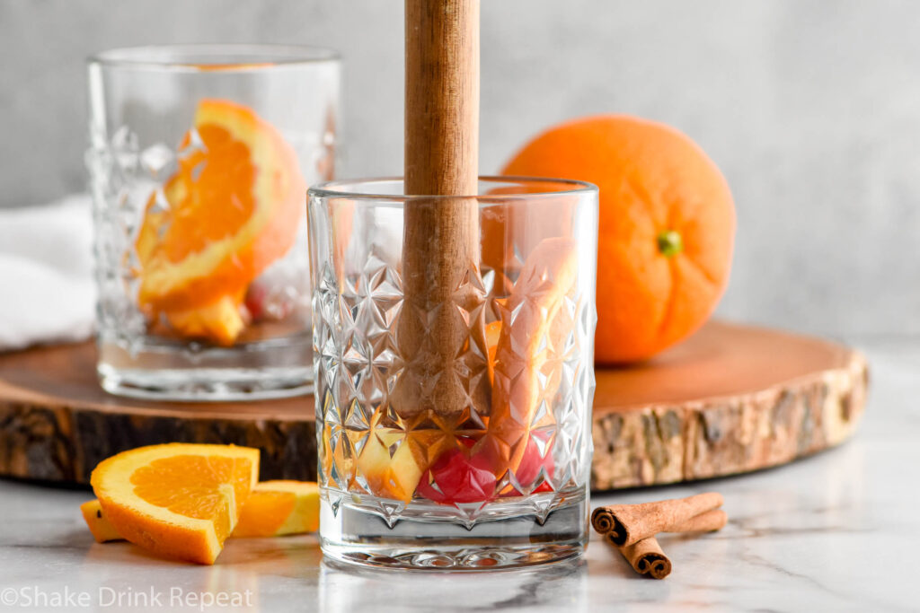 muddling autumn old fashioned ingredients in a glass with orange slices and cinnamon sticks sitting beside.