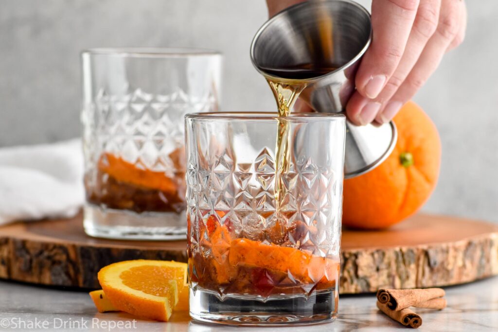 man's hand pouring bourbon into a glass of fall spiced old fashioned ingredients. Orange slices and cinnamon sticks sitting beside.