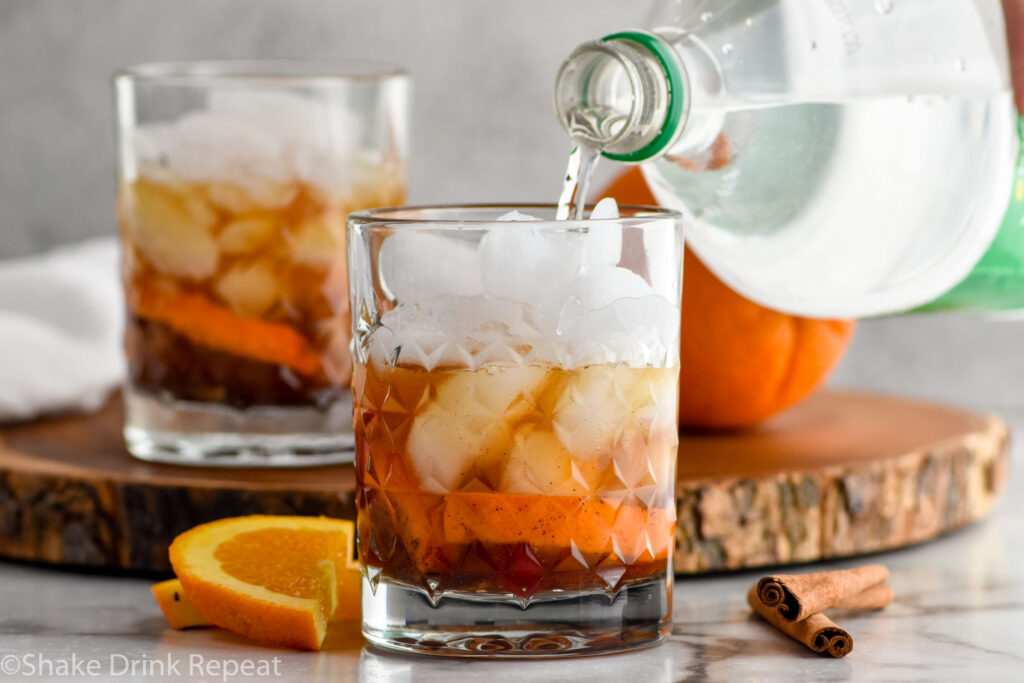 bottle of lemon lime soda pouring into a glass of fall spiced old fashioned ingredients with ice. Orange slices and cinnamon sticks sitting beside.