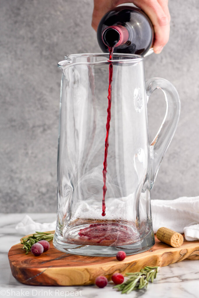 man's hand pouring a bottle of red wine into a glass pitcher to make friendsgiving sangria. Cranberries and rosemary sit beside to garnish