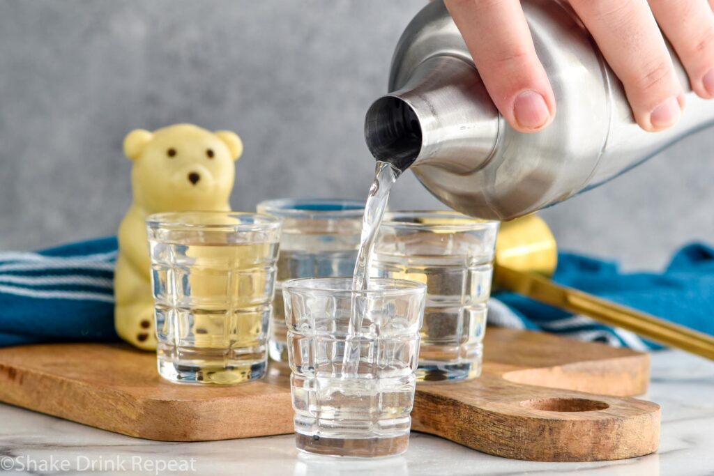 man's hand pouring cocktail shaker of polar bear shot ingredients into shot glass. Polar bear shots and polar bear garnish sit in background