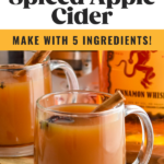 Pinterest graphic for crockpot spiced apple cider. Text says "crockpot apple cider so easy! make with 5 ingredients! shakedrinkrepeat.com" Image shows two mugs of crockpot spiced apple cider garnished with star anise and cinnamon sticks with bottle of fireball whiskey in background