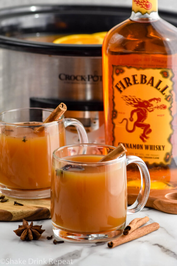 two mugs of spiced apple cider garnished with star anise and cinnamon sticks, bottle of fireball whiskey and crockpot sit in background
