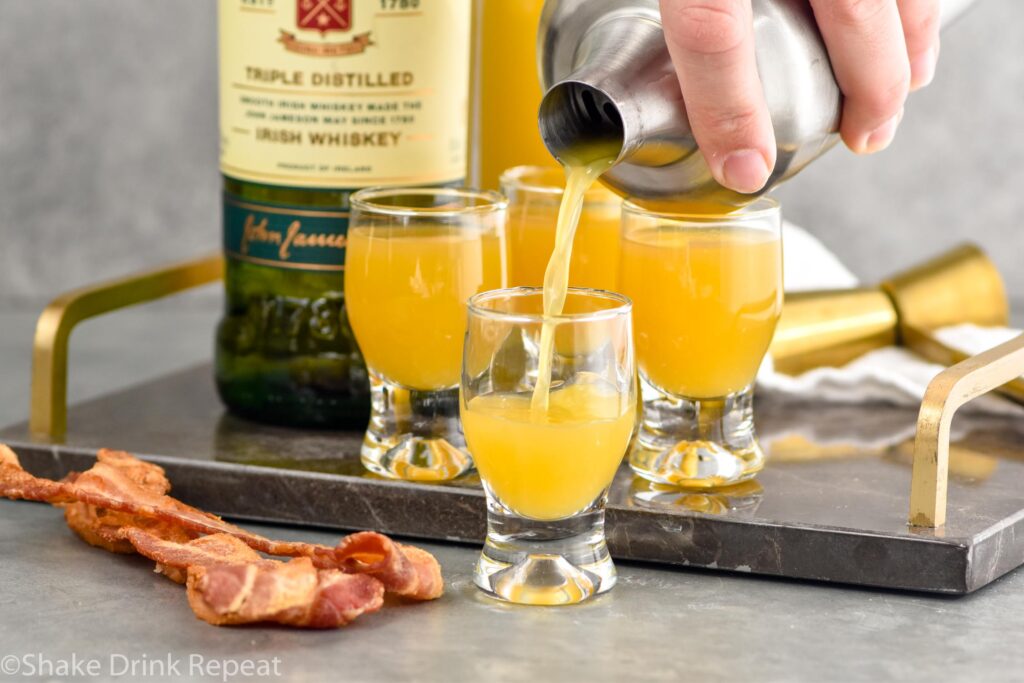 man's hand pouring cocktail shaker of breakfast shot ingredients into a shot glass. Strips of bacon, bottle of Jameson, and three shot glasses of breakfast shots sitting in background