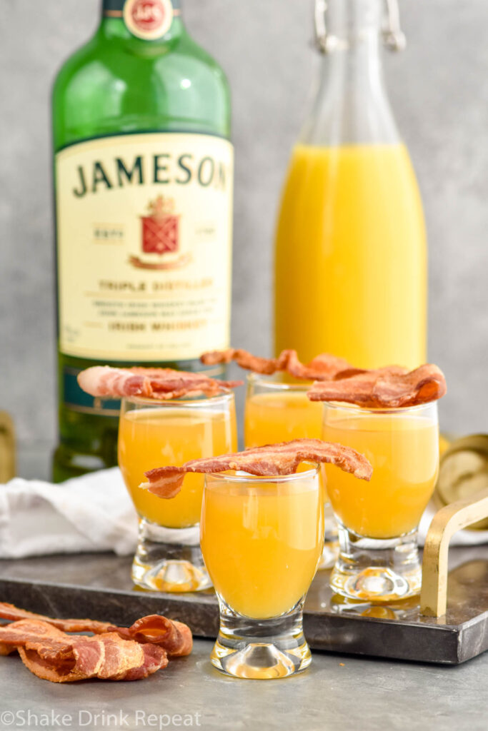 four shot glasses of breakfast shots with bacon, bottle of Jameson, and orange juice sitting in background