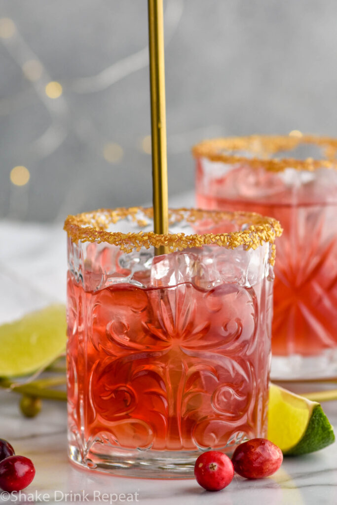 Front facing photo of a glass of cranberry margarita being stirred with a long golden spoon with a golden sugar rim and cranberries floating in the drink and around the glass.