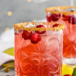 Front facing photo of a glass of cranberry margarita with a golden sugar rim and cranberries floating in the drink and around the glass with a second drink in the background