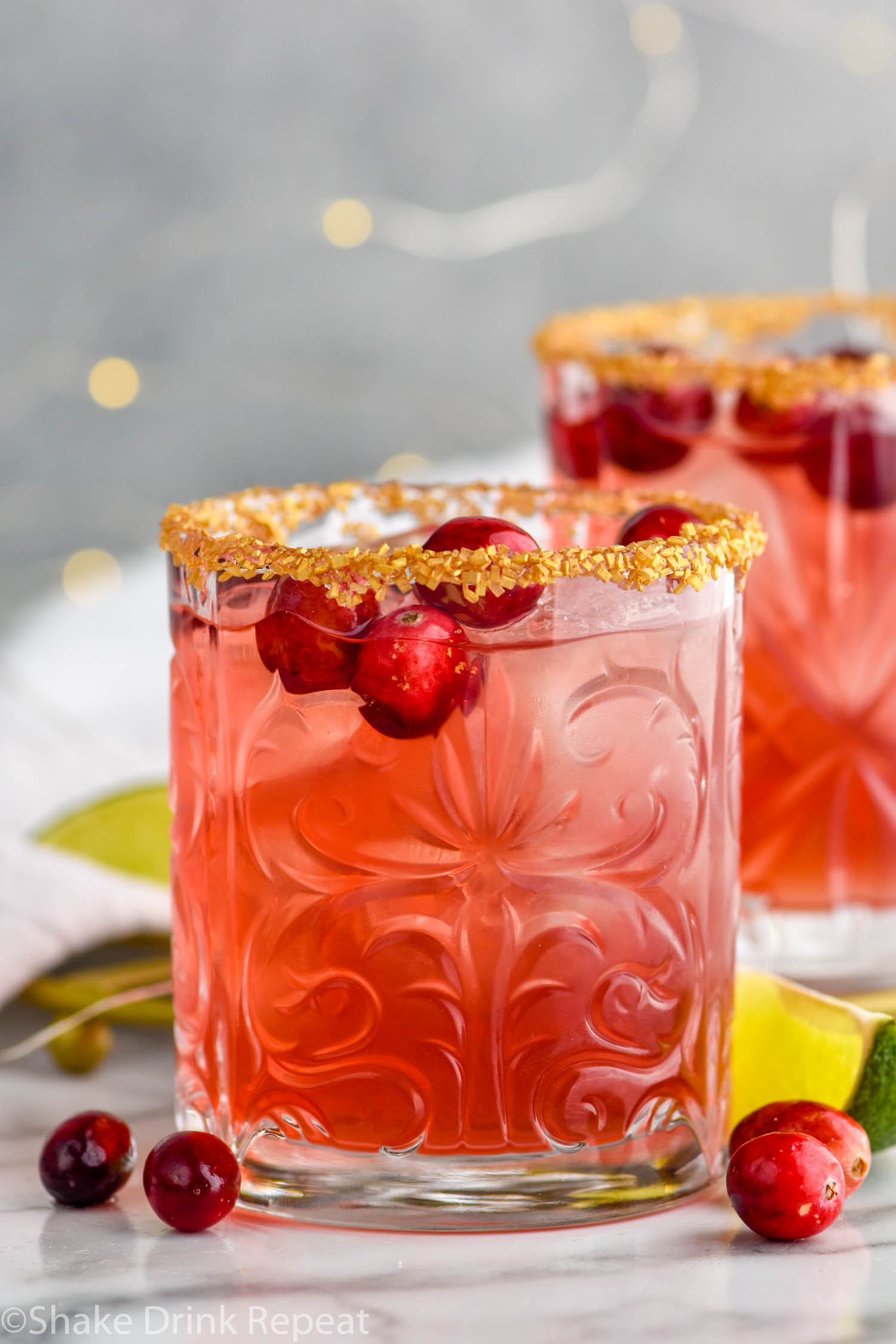 Front facing photo of a glass of cranberry margarita with a golden sugar rim and cranberries floating in the drink and around the glass with a second drink in the background