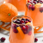 Photo of Cranberry Orange Margarita Sangria in a stemless wine glass with a golden rim with a second glass, pitcher, and orange in the background.