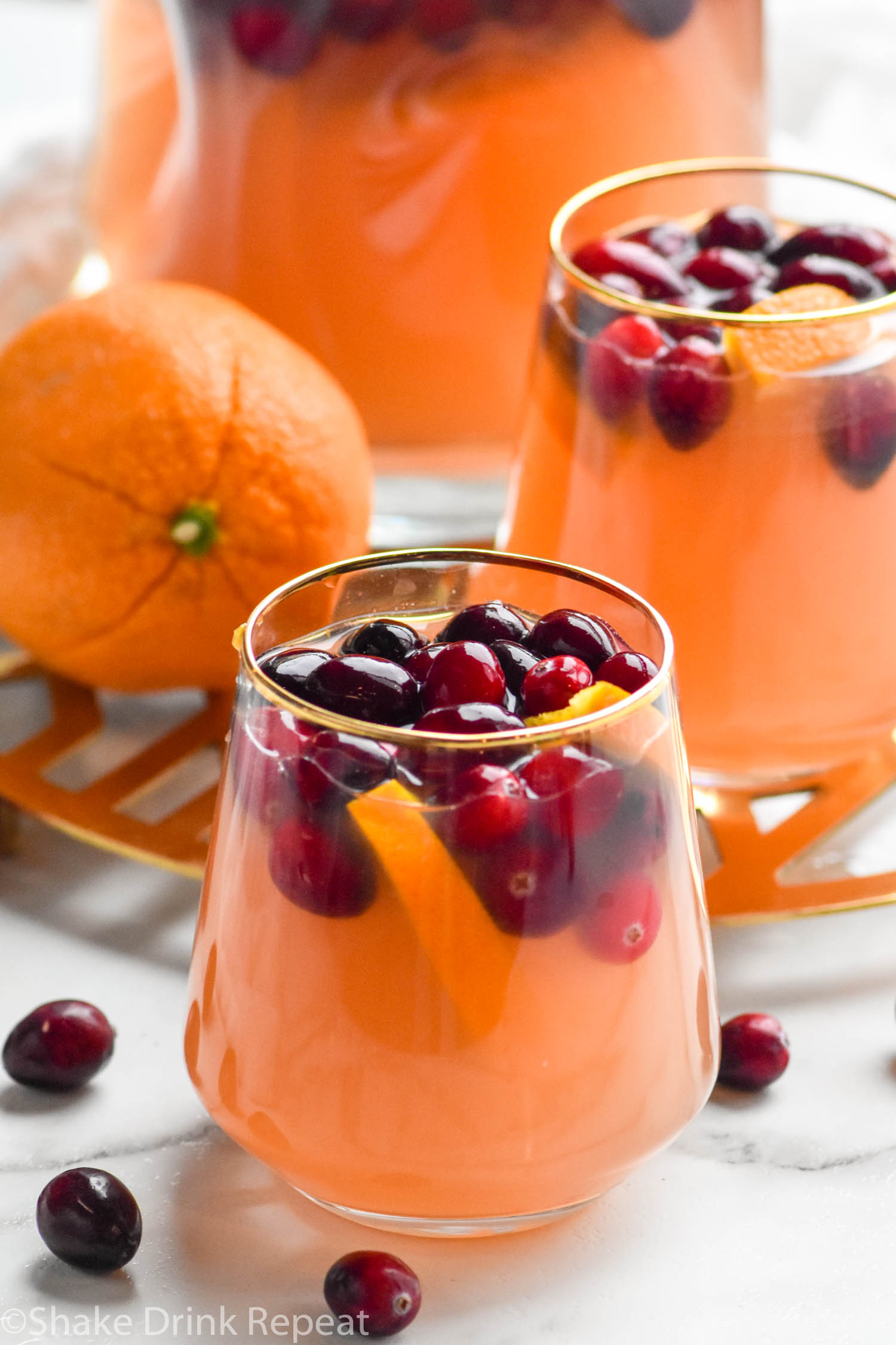 Photo of Cranberry Orange Margarita Sangria in a stemless wine glass with a golden rim with a second glass, pitcher, and orange in the background.