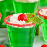 Close up front-facing photo of Drunken Elf Jello Shots topped with whipped cream, red hot, and mint, with mint and red hots on the table around the shots.