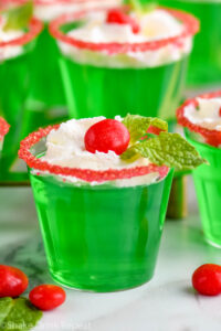 Close up front-facing photo of Drunken Elf Jello Shots topped with whipped cream, red hot, and mint, with mint and red hots on the table around the shots.