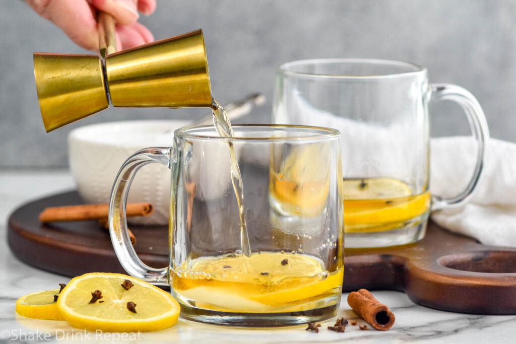 man's hand pouring jigger of bourbon into a mug with a lemon slice and cloves to make a hot toddy cocktail. Extra lemon slices with cloves and cinnamon sticks sitting beside, mug of hot toddy ingredients sitting in background