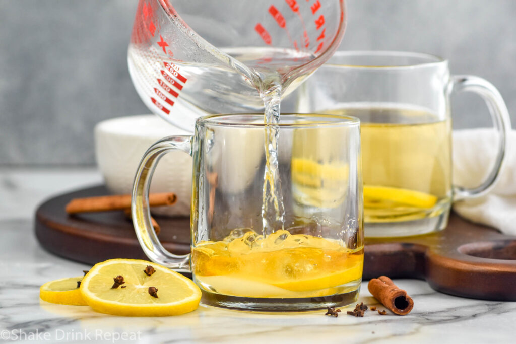 Measuring cup of hot water pouring into a mug of hot toddy ingredients. Lemon slices with cloves and cinnamon sticks sitting beside