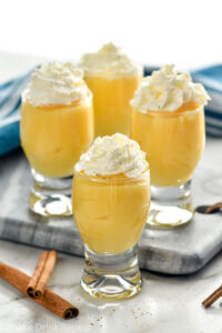 Overhead photo of RumChata Pudding Shots garnished with whipped cream. Cinnamon sticks lay on counter beside pudding shots.