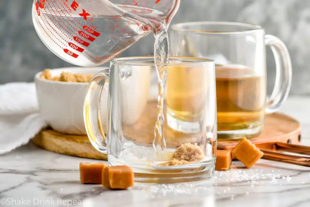 measuring cup of hot water pouring into a mug of brown sugar and salt to make a salted caramel hot buttered rum. Mug of hot buttered rum ingredients and bowl of brown sugar sitting in background and caramel candies sitting beside