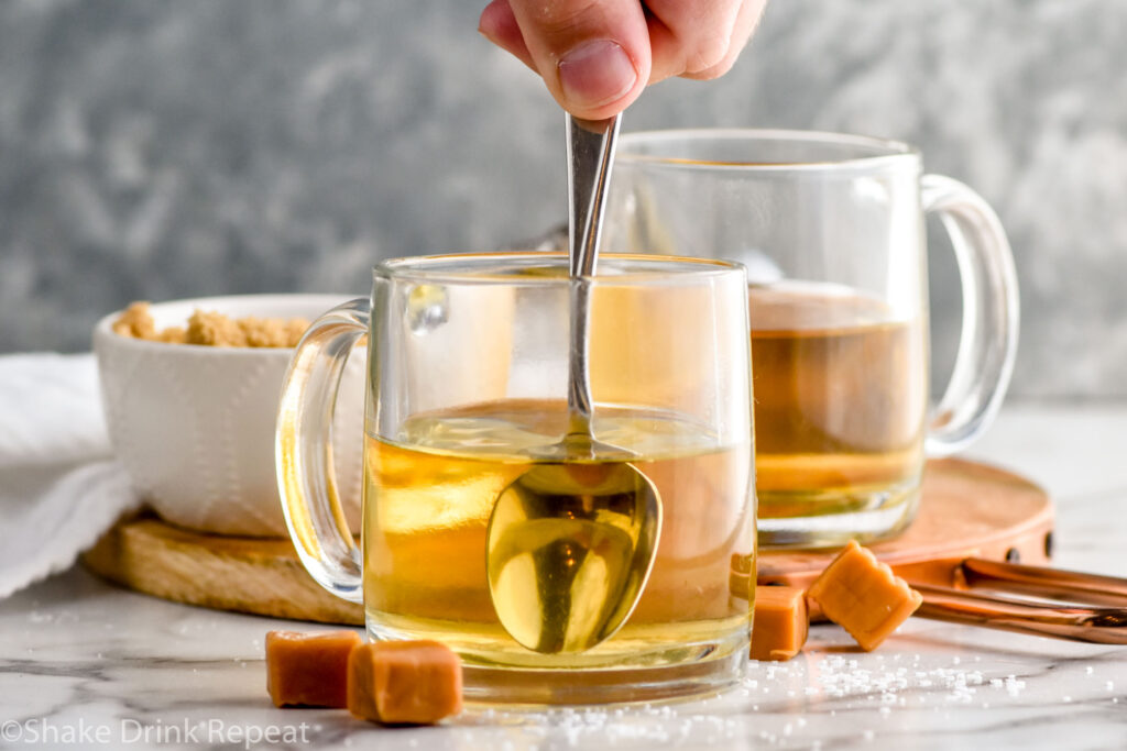 man's hand stirring mug of salted caramel buttered rum ingredients with second mug and bowl of brown sugar sitting in background and caramel candies sitting beside
